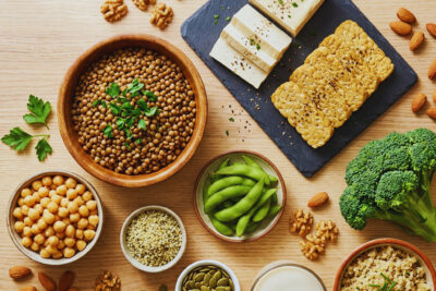 Vegan protein sources spread — lentils, tofu, tempeh, edamame, hemp seeds, almonds, and quinoa arranged on a wooden table